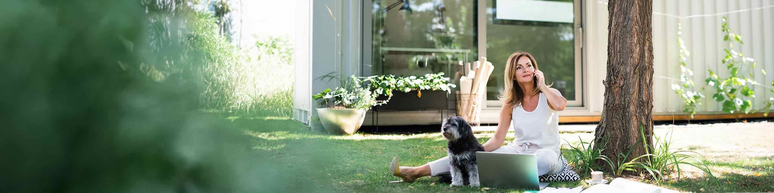 Frau mit Hund und Laptop im Garten telefoniert.
