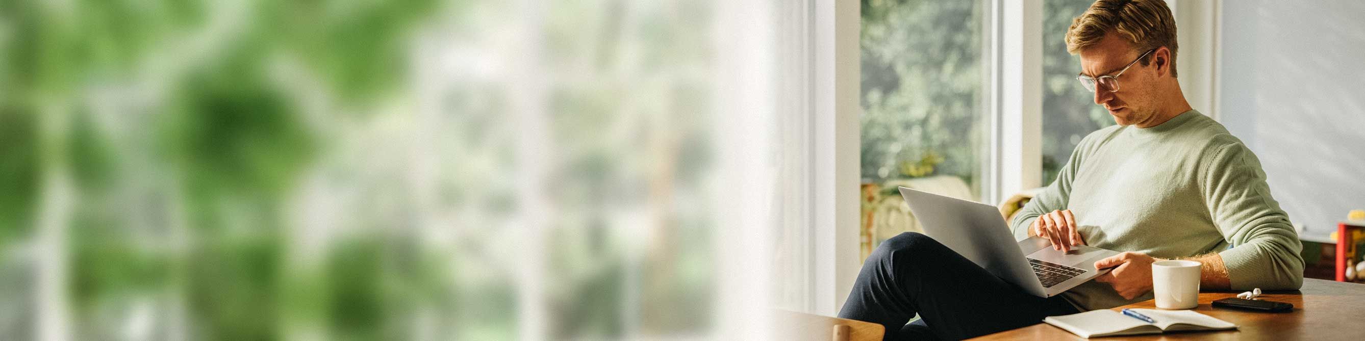 Mann auf Stuhl am Fenster schaut auf den Laptop auf seinem Schoß.