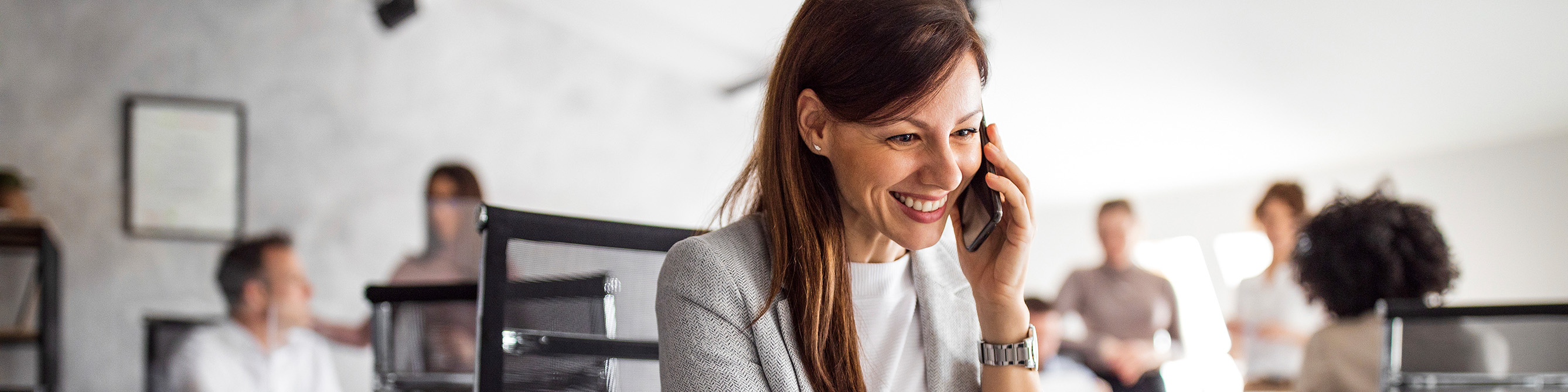 Eine Frau sitz in einem Büro und telefoniert mit einem Handy, sie lächelt in die Kamera.