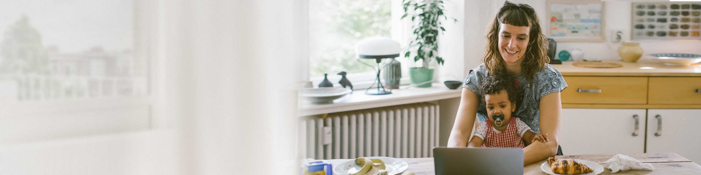 Frau sitzt am Tisch vor ihrem Laptop mit ihrem Kind auf dem Schoß.