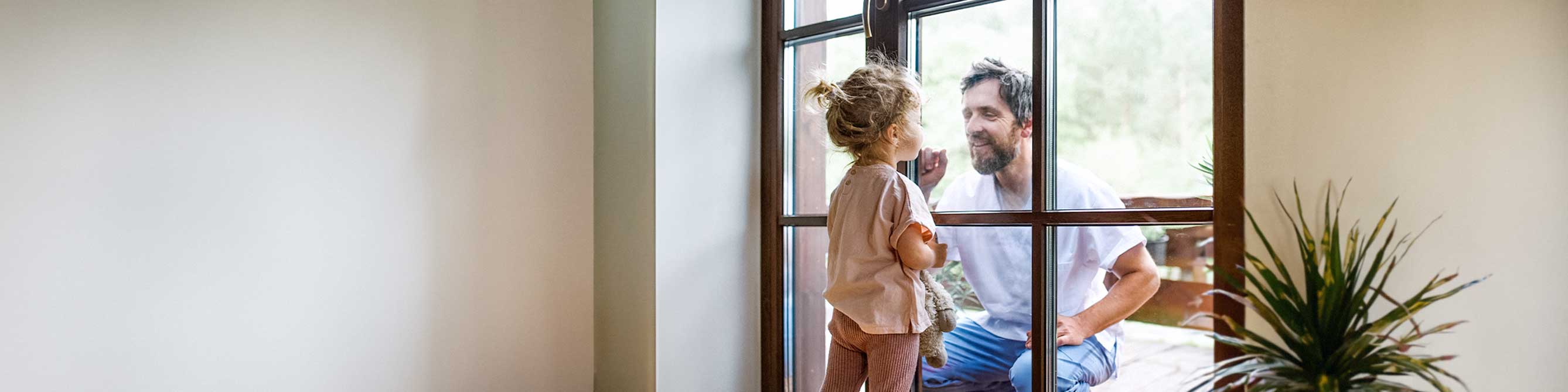 Vater und kleine Tochter schauen sich durch Fensterscheibe an und lächeln.