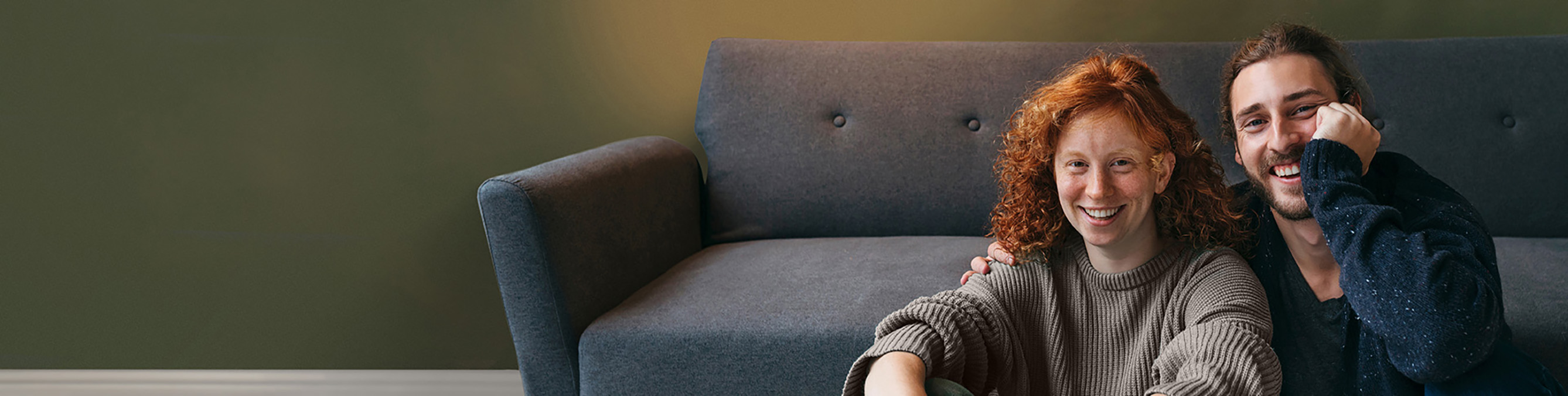 Ein junges paar sitzt vor einer Couch und lacht in die Kamera.