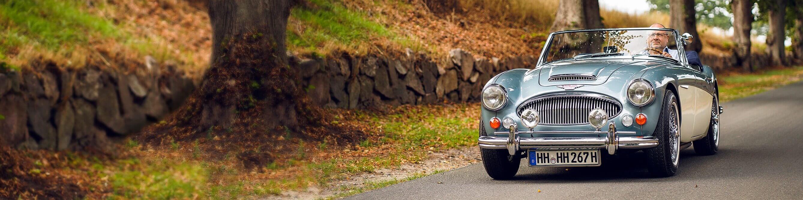 Klassischer hellblauer Oldtimer-Cabriolet mit einem Mann am Steuer und offenem Verdeck fährt auf einer Landstraße entlang einer Steinmauer im Grünen.