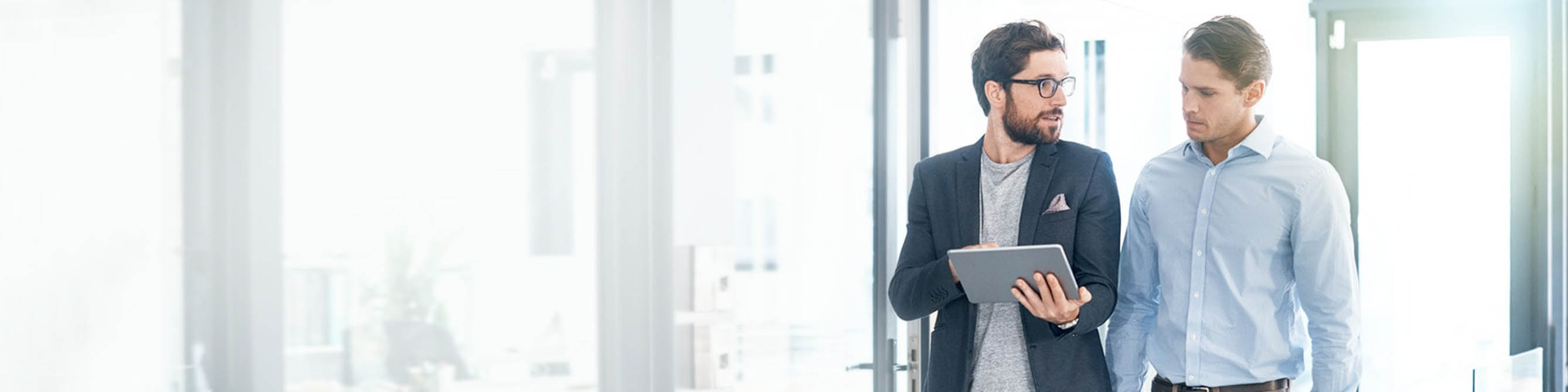 Zwei Männer stehen in einem Büro, einer hält ein Tablet in der Hand, beide unterhalten sich.