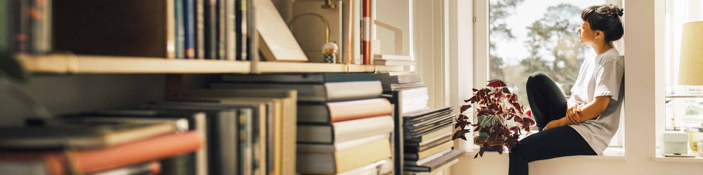 Junge Frau sitzt im Fensterrahmen und schaut hinaus, im Vordergrund sieht man ihr Bücherregal.