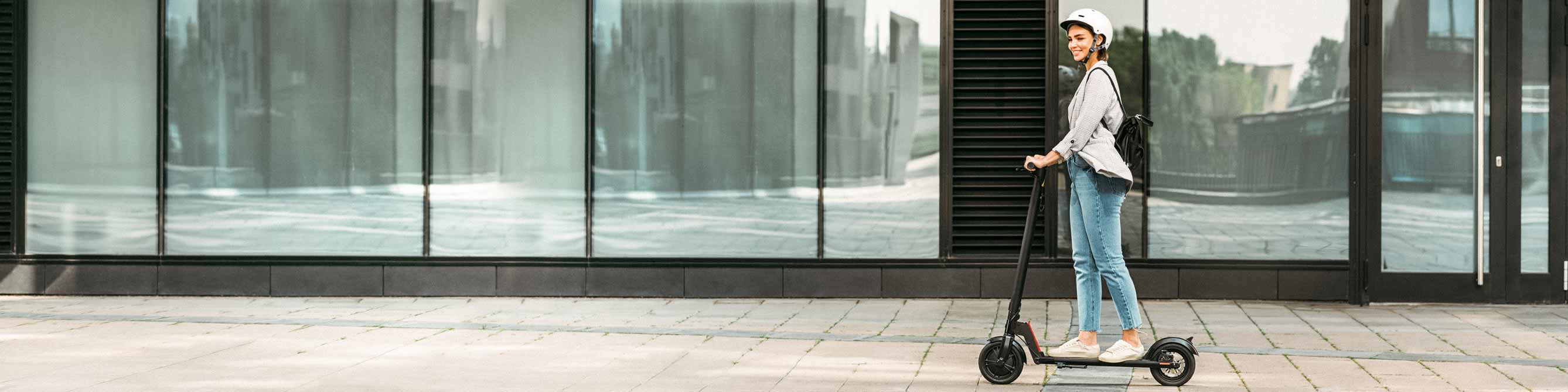 E-Scooter-Versicherung: Frau fährt auf einem E-Scooter entlang einer Fensterfront