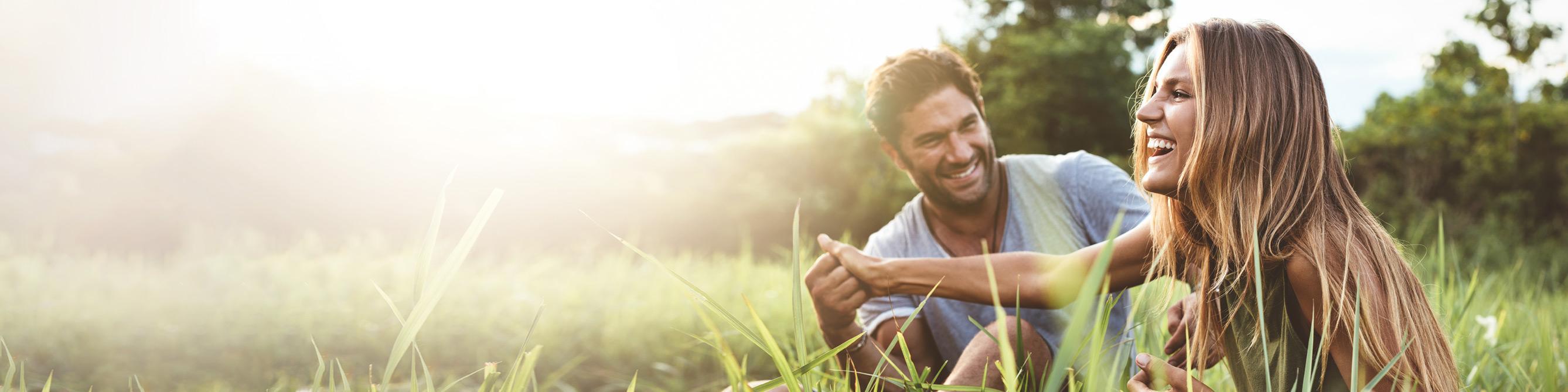 Ein junger Mann und eine junge Frau sitzen in der Abendsonne auf einer Wiese und lachen sich an.
