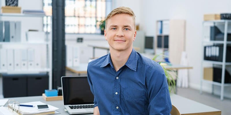 Ein junger Mann mit blauem Hemd sitzt im Büro und schaut in die Kamera.