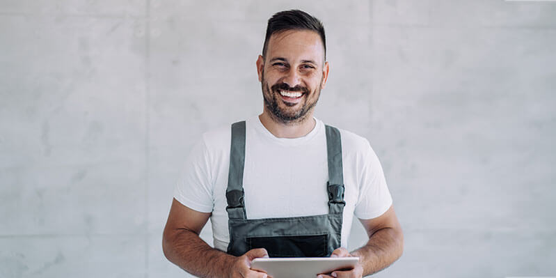 Ein junger Mann mit Latzhose steht vor einer hellen Wand und bedient ein Tablet