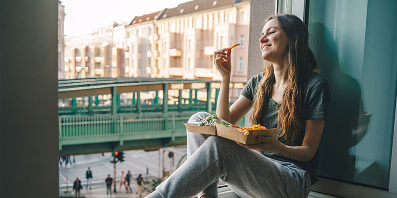 Eine junge Frau sitzt auf einer Fensterbrüstung und isst Pommes, während im Hintergrund die Stadt pulsiert.