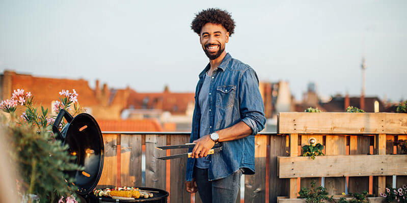 Ein junger Mann steht lachend auf einem Balkon und grillt