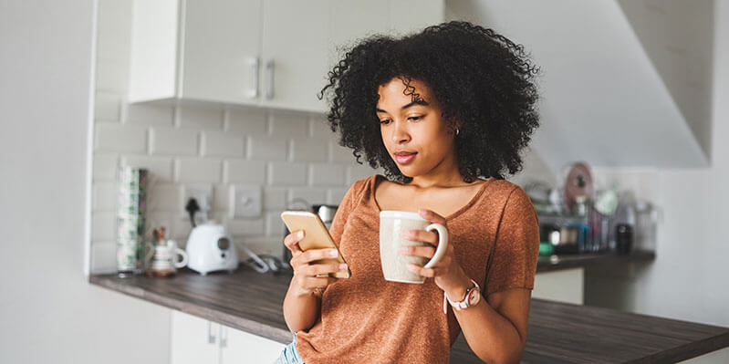 Eine junge Frau steht in der Küche, trinkt einen Kaffee und schaut auf ihr Handy