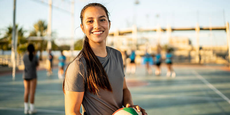 Eine junge Frau steht lachend auf einem Sportplatz und hält einen Volleyball in der Hand 