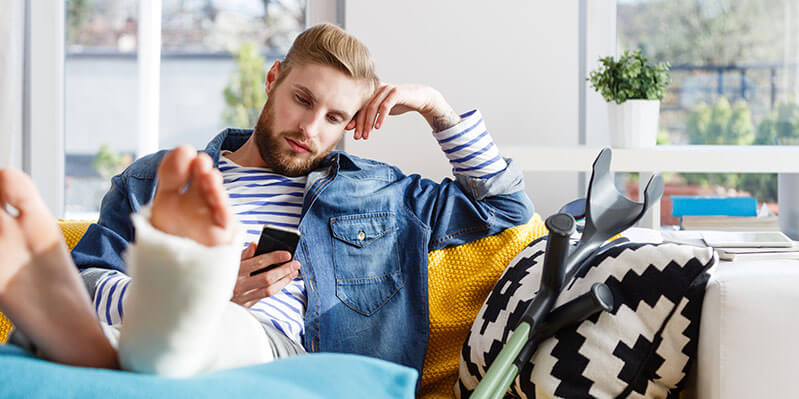 Ein junger Mann mit gebrochenem Bein sitzt auf einer Couch und schaut aufs Handy