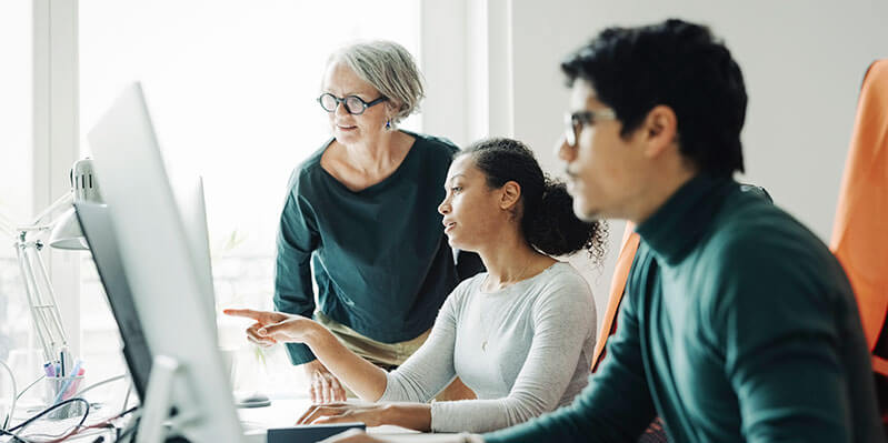 Eine Gruppe von zwei Frauen und einem Mann sitzen in einem Büro und schauen auf zwei Computer 